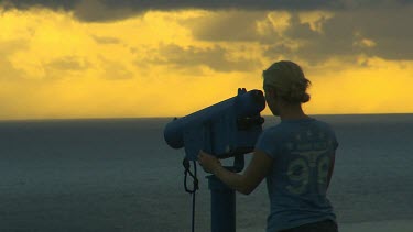 Young woman in shadow looking through telescope out to sea. Sunset with yellow golden sky and small puffy cumulus clouds.