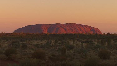 Uluru, red sunset reflecting off rock. Desert plains in foreground in shadow.