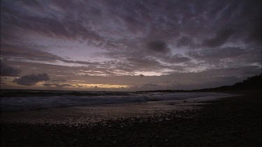 Wide Shot. Pebble beach with rocks in evening light. Sea and sky. Sun set in distance, horizon. Clouds float overhead, blown in gentle wind breeze.