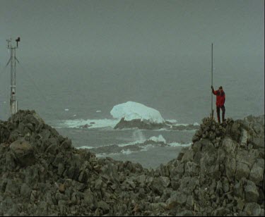 Man on Rocky Outcrop Proclamation Hill, Antarctiva. Iceberg in background. It is snowing.