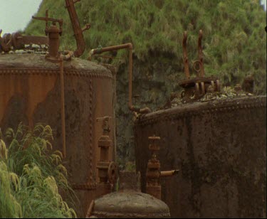 Still shot of rusty industrial equipment, possibly tanks, slowly deacying on the edge of the bay