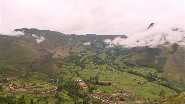 Pan across a valley and a town to a mountain side village ruins in Peru