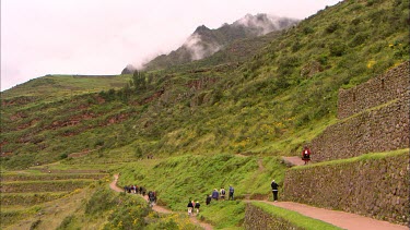 Tourist walking across rock terraces in Peru, cloud covered peaks in the background