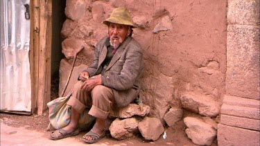 Old Peruvian man sitting on a pile of rocks leaning against a terracotta coloured wall