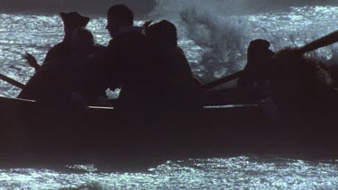 Historical long boat coming to shore through crashing waves. People in silhouette,