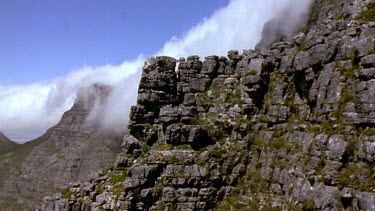 Track up along side of mountain, clouds tumbling down to cover the cliff face.