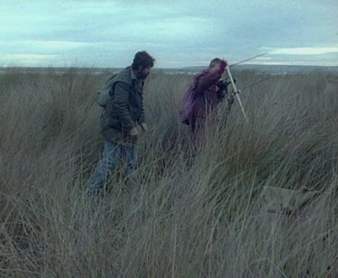 Evening. Low light. Scientists carrying radio transmission and receiving equipment walk through long grass.