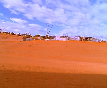 Past outback shacks corrugated iron settlement in middle of red earth desert.