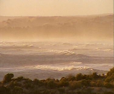 Sunset. Rough ocean storm waves and surf. Sand dunes in background. The air is filled with the fine dust of the desert and ocean spray.