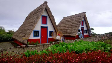 Local Lady Lets Tourist Try On Hat At Traditional Thatched Triangular Houses, Santana, Madeira, Portugal